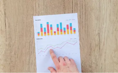 A hand pointing at a piece of paper with colorful bar and line graphs, placed on a wooden surface.